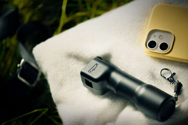 Plegium zelfverdedigingsspray in de kleur zwart liggend op een beige handdoek naast een gele smartphone